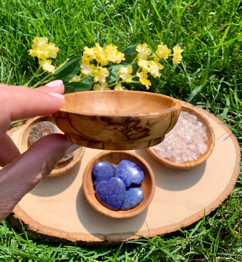 K&ouml;nnte beinhalten: Vier kleine Holzschalen mit verschiedenen Kristallen. Die Schalen sind auf einer Holzscheibe angeordnet. Die Schalen enthalten blaue herzf&ouml;rmige Steine, rosa Kristalle und eine versteinerte Muschel.