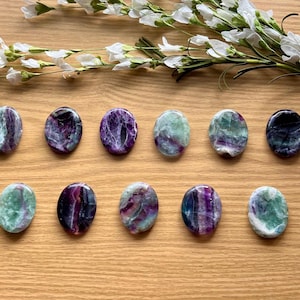 May include: A collection of oval-shaped fluorite stones in shades of purple, green, and white, arranged on a wooden surface. White flowers are placed at the top of the image. The stones have a polished, smooth texture.