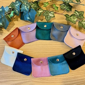 May include: A collection of small, square velvet pouches in various colors, including orange, purple, green, gray, pink, white, blue, and black. Each pouch has a rounded flap secured with a gold-colored snap. Green leafy vines are in the background.
