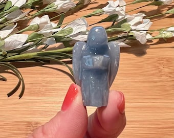 May include: A small, light blue angel figurine with outstretched wings. The angel is carved from a translucent material and held between fingers. White flowers and green stems are in the background.