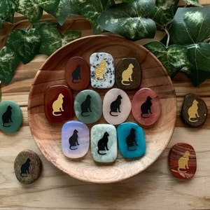 May include: A collection of decorative stones, each featuring a cat silhouette. The stones vary in color, including green, red, and blue, and are arranged in a wooden bowl. Some cats are gold, others black.