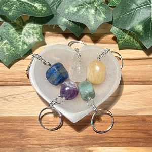 May include: A set of seven chakra crystal keychains in a white heart-shaped dish. The crystals are clear quartz, amethyst, green aventurine, citrine, lapis lazuli, and red jasper. The keychains are silver.