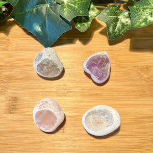 May include: Four polished gemstones in shades of clear, purple, pink, and white, arranged on a wooden surface. The stones have a rough, natural exterior and a smooth, polished interior. Green leaves are in the background.