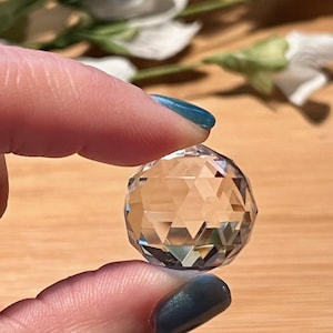 May include: A clear, faceted crystal ball held between two fingers. The sphere reflects light, creating a sparkling effect. The background includes a wooden surface and white flowers.