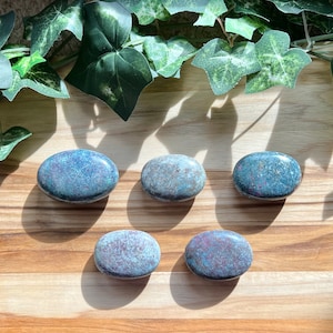 May include: Five smooth, oval-shaped stones with a mottled blue and green color. The stones are arranged in a row on a wooden surface.
