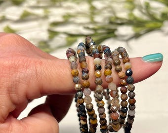 May include: A multi-strand bracelet composed of small, faceted, round beads in earthy tones including brown, tan, gray, blue, and yellow. The beads are strung on a flexible wire, creating a layered appearance with a reflective, textured surface.