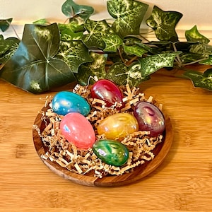May include: Six iridescent gemstone eggs in a wooden bowl with shredded paper. The eggs are in shades of pink, blue, green, yellow, and red.