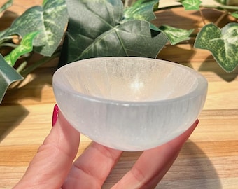 May include: A small, round, white selenite bowl. The bowl is held in a hand with red nail polish. Green leaves and a wooden surface are in the background. The bowl has a smooth, translucent appearance.