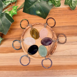 May include: Six oval-shaped gemstone keychains arranged in a circle on a wooden bowl. The stones include tiger's eye, rose quartz, amethyst, black obsidian, light green aventurine, and clear quartz, each attached to a silver-toned metal ring.