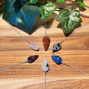 May include: A collection of gemstone pendulums on silver chains arranged in a circular pattern. The gemstones are various colors, including pink, red, blue, and black, and are displayed on a wooden surface. Green leaves are in the background.
