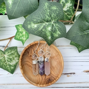 K&ouml;nnte beinhalten: Drei Kristallspitzen-Anh&auml;nger mit silbernen Kappen und klaren Kristallkugeln. Die Anh&auml;nger liegen auf einer Holzoberfl&auml;che und sind in einer Reihe angeordnet. Die Kristalle sind klar, Amethyst und Rosenquarz.