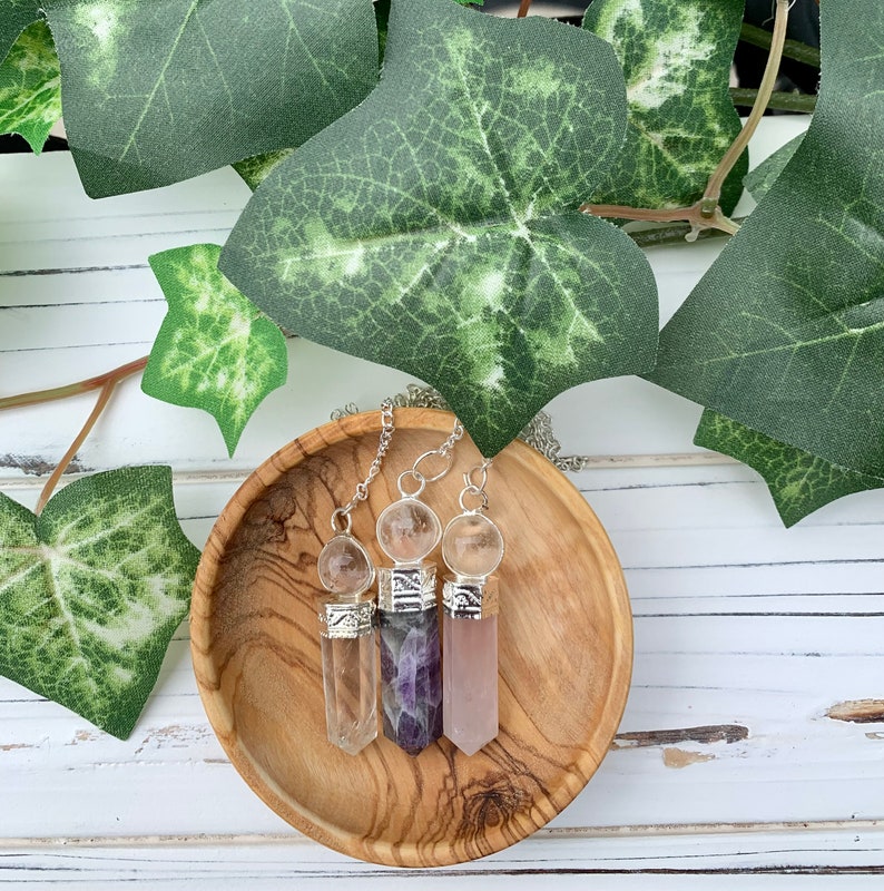 K&ouml;nnte beinhalten: Drei Kristallspitzen-Anh&auml;nger mit silbernen Kappen und klaren Kristallkugeln. Die Anh&auml;nger liegen auf einer Holzoberfl&auml;che und sind in einer Reihe angeordnet. Die Kristalle sind klar, Amethyst und Rosenquarz.