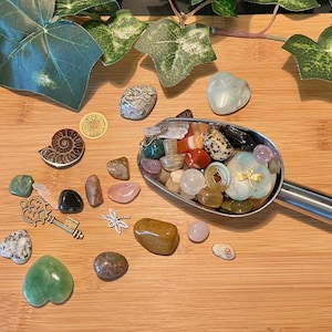 May include: A metal scoop filled with a variety of colorful stones, crystals, and charms. The scoop is on a wooden surface with other stones and charms scattered around it.