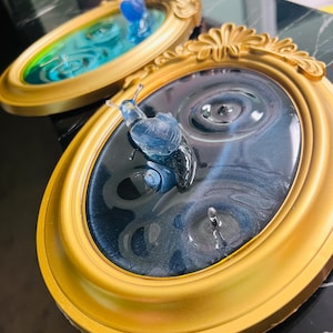 以下が含まれることがあります： 金色のフレームに入った2つの楕円形の樹脂アート作品。1つは青いカタツムリと濃い青色の渦巻き模様、もう1つは青と緑の渦巻き模様です。フレームには装飾的なディテールがあります。