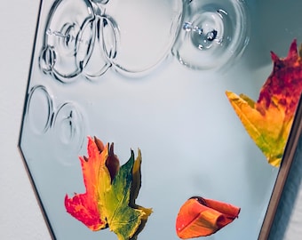 May include: A hexagonal mirror with a bronze frame. The mirror's surface features a clear, textured design of overlapping circles. Colorful autumn leaves in shades of red, yellow, and green are reflected in the mirror.