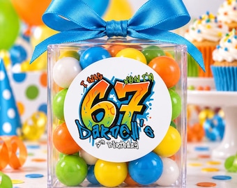May include: A clear square plastic box filled with colorful gumballs, topped with a bright blue satin ribbon bow. A circular white label on the front features the text "I WAS NOW I'M 67 DARNELL'S 7th BIRTHDAY" in a bold, graffiti-style font.