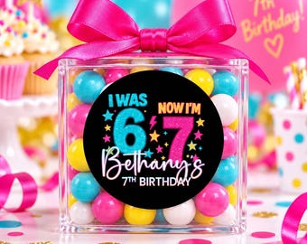 May include: A clear square plastic container filled with colorful round candies, topped with a bright pink satin ribbon bow. A circular black label on the front features the text I WAS 6 NOW I'M 7, Bethany's 7TH BIRTHDAY, with colorful stars.
