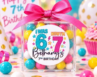May include: A clear square plastic container filled with colorful gumballs, topped with a bright pink satin ribbon bow. A circular label on the front features the text I WAS 6 NOW I'M 7, Bethany's 7TH BIRTHDAY, decorated with colorful stars.