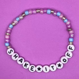 May include: A white beaded bracelet with the words "Shake It Off" spelled out in black letters. The bracelet is made with small, colorful beads in shades of pink, blue, and silver.