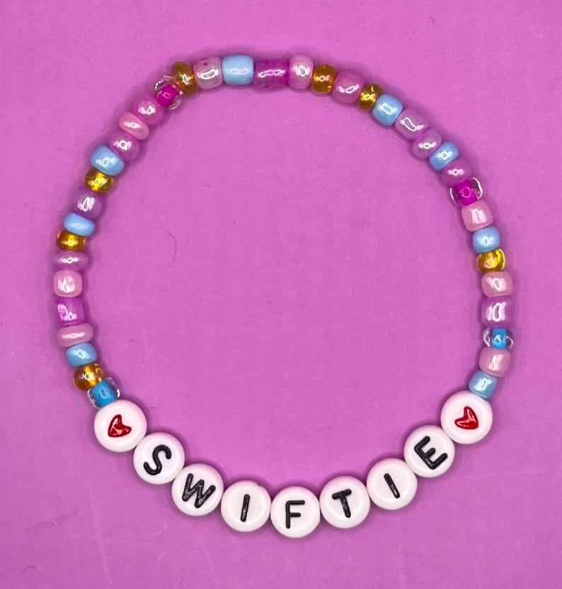 Puede incluir: Un brazalete de cuentas con cuentas de color rosa pastel, azul y dorado. El brazalete deletrea "SWIFTIE" con un coraz&oacute;n rojo a cada lado.