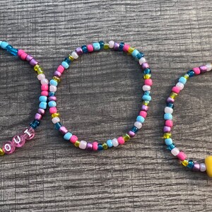 May include: Three circular beaded bracelets featuring a mix of small pink, blue, purple, yellow, and white glass beads. One bracelet includes white letter beads spelling WOLF OUT, and another features a single orange, yellow, and white candy corn charm.