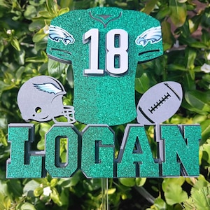 May include: A glittery green football-themed decoration featuring a jersey with the number 18, a football helmet, and a football. The name "LOGAN" is displayed in green glitter letters. The decoration is designed for a sports fan.