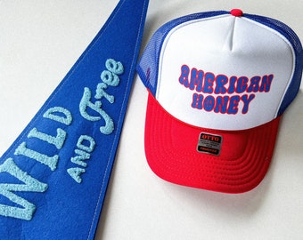 Puede incluir: Un banderín azul y blanco con el texto "Wild and Free" y una gorra de camionero roja y blanca con el texto "American Honey" y una etiqueta en blanco y negro que dice "Otto Trucker".