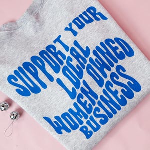 May include: A heather gray sweatshirt with the text "SUPPORT YOUR LOCAL OWNED WOMEN BUSINESS" in blue, with two small silver disco ball ornaments. The sweatshirt is folded on a pink surface.