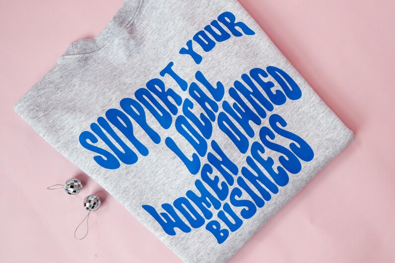 May include: A heather gray sweatshirt with the text "SUPPORT YOUR LOCAL OWNED WOMEN BUSINESS" in blue, with two small silver disco ball ornaments. The sweatshirt is folded on a pink surface.
