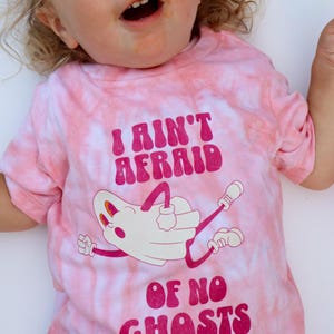 Puede incluir: Camiseta rosa te&ntilde;ida con la frase "I AIN'T AFRAID OF NO GHOSTS" en magenta. La camiseta presenta una ilustraci&oacute;n de fantasma de dibujos animados en blanco con detalles magenta. La camiseta es de manga corta.