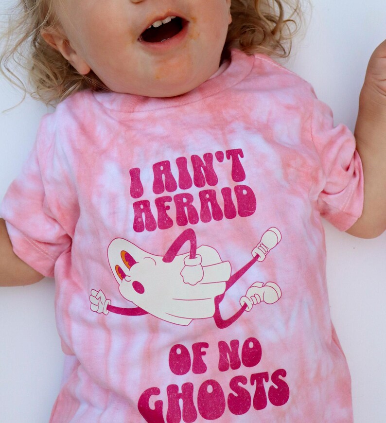 Puede incluir: Camiseta rosa te&ntilde;ida con la frase "I AIN'T AFRAID OF NO GHOSTS" en magenta. La camiseta presenta una ilustraci&oacute;n de fantasma de dibujos animados en blanco con detalles magenta. La camiseta es de manga corta.