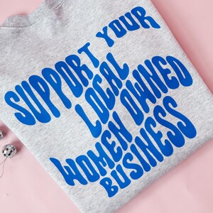 May include: A heather gray sweatshirt with the blue text "SUPPORT YOUR LOCAL OWNED WOMEN BUSINESS". Two small silver disco ball ornaments are on a pink background.
