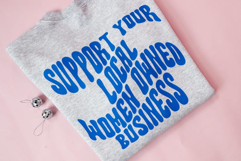 May include: A heather gray sweatshirt with the blue text "SUPPORT YOUR LOCAL OWNED WOMEN BUSINESS". Two small silver disco ball ornaments are on a pink background.