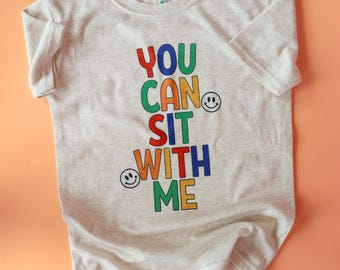 Puede incluir: Una camiseta gris claro con el texto "YOU CAN SIT WITH ME" en letras de colores y gráficos de caras sonrientes. La camiseta está hecha de un material suave y tiene mangas cortas. El fondo es de color naranja liso.