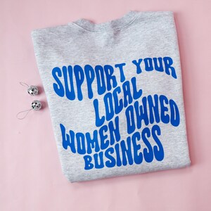 May include: A gray sweatshirt with the blue text "SUPPORT YOUR LOCAL OWNED WOMEN BUSINESS" is displayed on a pink surface. Two small silver disco ball ornaments are placed to the left of the sweatshirt.