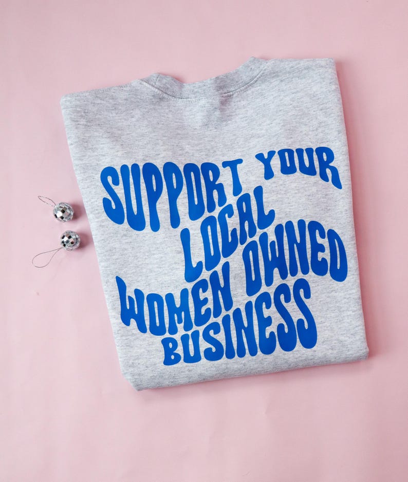 May include: A gray sweatshirt with the blue text "SUPPORT YOUR LOCAL OWNED WOMEN BUSINESS" is displayed on a pink surface. Two small silver disco ball ornaments are placed to the left of the sweatshirt.