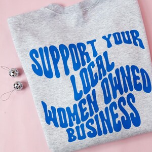 May include: A heather gray sweatshirt with the blue text "SUPPORT YOUR LOCAL OWNED WOMEN BUSINESS". Two small silver disco ball ornaments are on the left side of the sweatshirt. The background is pink.