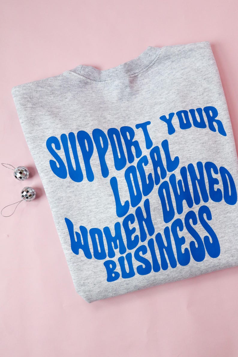 May include: A heather gray sweatshirt with the blue text "SUPPORT YOUR LOCAL OWNED WOMEN BUSINESS". Two small silver disco ball ornaments are on the left side of the sweatshirt. The background is pink.