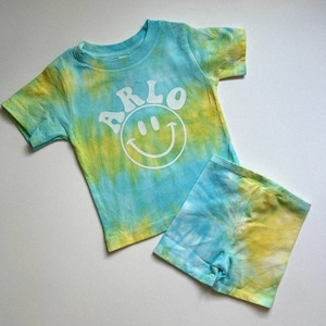 May include: A blue and yellow tie-dye t-shirt and shorts set with a white smiley face and the name "ARLO" printed on the shirt.