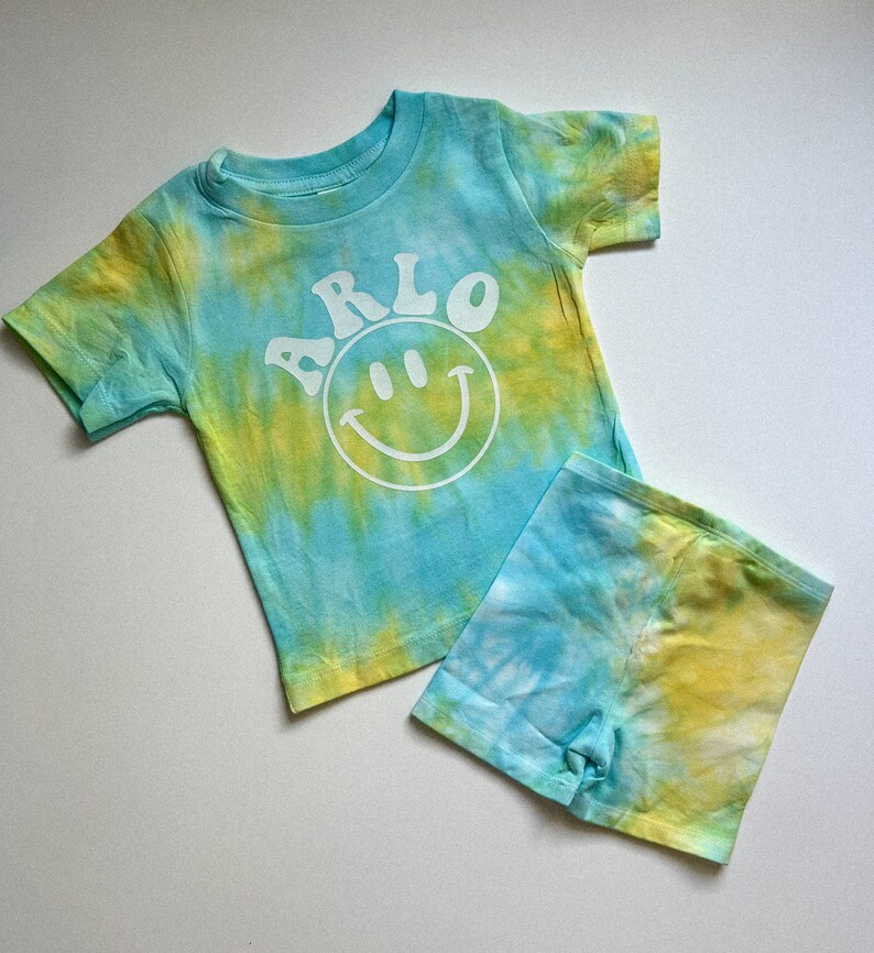 May include: A blue and yellow tie-dye t-shirt and shorts set with a white smiley face and the name "ARLO" printed on the shirt.
