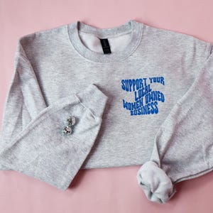 May include: A light gray sweatshirt with the text "SUPPORT YOUR LOCAL OWNED WOMEN BUSINESS" in blue. The sweatshirt has long sleeves and a crew neck. Two silver disco ball earrings are on the left sleeve.