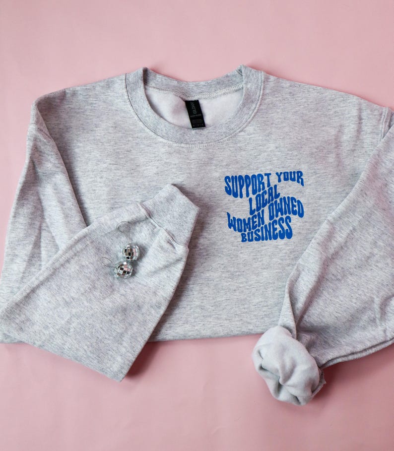May include: A light gray sweatshirt with the text "SUPPORT YOUR LOCAL OWNED WOMEN BUSINESS" in blue. The sweatshirt has long sleeves and a crew neck. Two silver disco ball earrings are on the left sleeve.