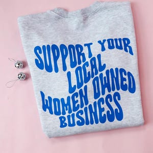May include: A heather gray sweatshirt with the blue text "SUPPORT YOUR LOCAL OWNED WOMEN BUSINESS." Two small silver disco ball ornaments are on the left side of the sweatshirt. The background is a light pink.