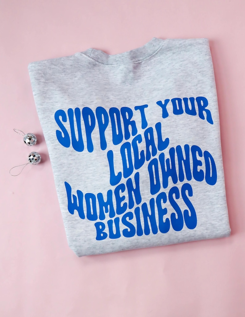 May include: A heather gray sweatshirt with the blue text "SUPPORT YOUR LOCAL OWNED WOMEN BUSINESS." Two small silver disco ball ornaments are on the left side of the sweatshirt. The background is a light pink.