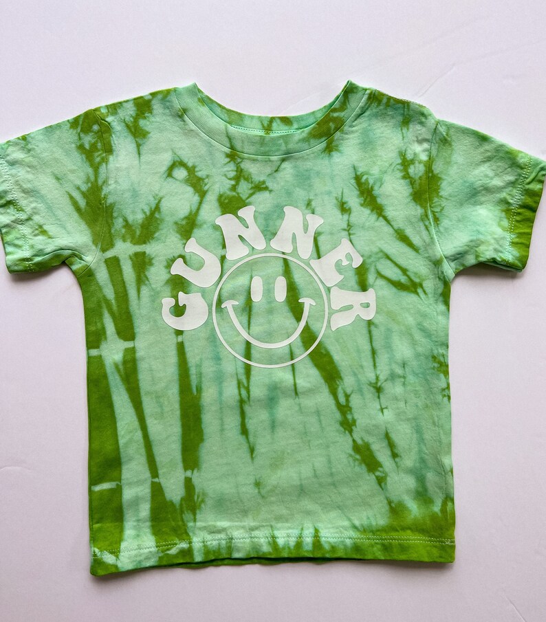 May include: Green tie-dye t-shirt with a white smiley face and the word "GUNNER" in a retro font.