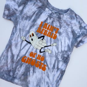 Puede incluir: Una camiseta gris y blanca con estampado tie-dye con un fantasma de dibujos animados y el texto naranja "I Ain't Afraid Of No Ghosts". El fantasma es blanco con brazos y piernas negros. La camiseta es de cuello redondo y manga corta.