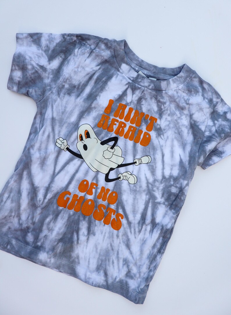 Puede incluir: Una camiseta gris y blanca con estampado tie-dye con un fantasma de dibujos animados y el texto naranja "I Ain't Afraid Of No Ghosts". El fantasma es blanco con brazos y piernas negros. La camiseta es de cuello redondo y manga corta.