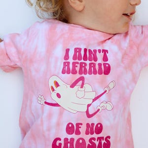 Puede incluir: Camiseta rosa te&ntilde;ida con la frase "I Ain't Afraid Of No Ghosts" en magenta. La camiseta presenta una ilustraci&oacute;n de un fantasma de dibujos animados con brazos y piernas. La camiseta es de material suave.