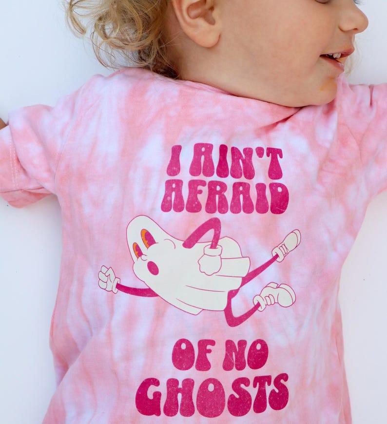 Puede incluir: Camiseta rosa te&ntilde;ida con la frase "I Ain't Afraid Of No Ghosts" en magenta. La camiseta presenta una ilustraci&oacute;n de un fantasma de dibujos animados con brazos y piernas. La camiseta es de material suave.