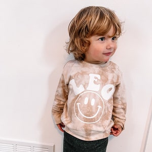 May include: A light brown tie-dye sweatshirt with a white smiley face and the name "LEO" in a retro font.